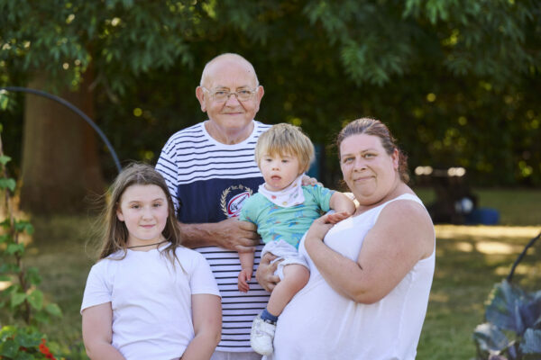 A young boy who has Downs syndrome with his grandad , sister and mum, outside in summer, smiling at the camera